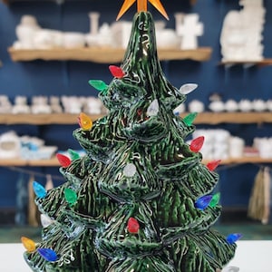 May include: A ceramic Christmas tree with a green glaze and a red base. The tree is decorated with colorful lights and a star on top.