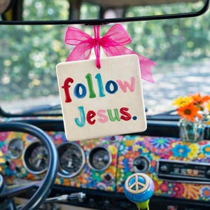 May include: A colorful interior of a vintage vehicle, featuring a dashboard with a floral pattern. A square sign with the words "follow Jesus" hangs from the rearview mirror, tied with a pink bow. A peace sign adorns the gear shift.
