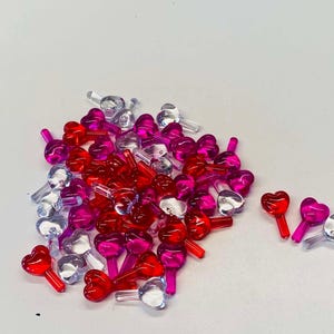 Może przedstawiać: Kolekcja przezroczystych, plastikowych osłon żarówek w kształcie serca w kolorze czerwonym, różowym i przezroczystym. Małe elementy dekoracyjne są rozrzucone na białej powierzchni, a kilka oddzielonych od głównej kupki.