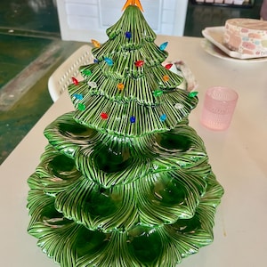 Könnte beinhalten: Ein Weihnachtsbaum aus Keramik mit glänzend grüner Oberfläche. Der Baum ist mit bunten Lichtern geschmückt und mit einem orangefarbenen Stern gekrönt. Der Sockel des Baumes hat ein mehrstufiges Design, ideal für Weihnachtsleckereien.