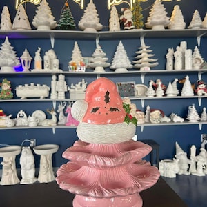 May include: A pink ceramic Christmas tree with a Santa Claus hat topper. The tree has three tiers and a round base. The background features shelves filled with various ceramic Christmas decorations, and a neon sign that reads "This must be the place."