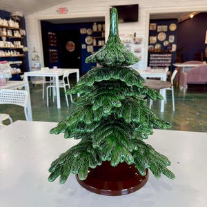 Puede incluir: Un árbol de Navidad de cerámica con un acabado verde brillante. El árbol tiene un diseño detallado y texturizado con una base marrón. El árbol se exhibe sobre una superficie blanca, con un fondo borroso de una habitación con mesas y sillas.
