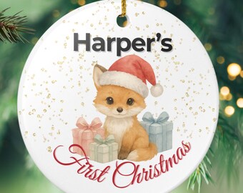May include: A white ceramic Christmas bauble featuring a fox in a Santa hat, the text "Harper's First Christmas", and three wrapped gifts. The bauble hangs from a gold string, set against a backdrop of Christmas tree foliage.