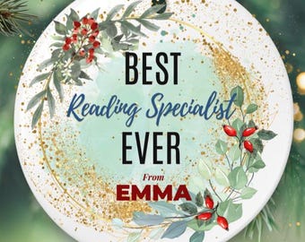 Puede incluir: Adorno navideño redondo de cerámica blanca con el texto "BEST Reading Specialist EVER From EMMA". El adorno tiene un borde dorado brillante y está decorado con hojas verdes y bayas rojas. Una cuerda dorada sirve para colgarlo, aprox. 8 cm de diámetro.