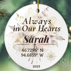 May include: A white ceramic Christmas ornament with the text "Always in Our Hearts Sarah" and coordinates. The ornament features a floral design in soft colors, a compass, and the year 2025. It hangs from a gold string.