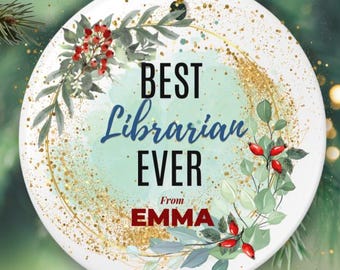 Puede incluir: Adorno navideño redondo y blanco con el texto "BEST LIBRARIAN EVER" en escritura negra y azul, con "From EMMA" en rojo. El adorno está decorado con hojas verdes, bayas rojas y detalles de purpurina dorada.