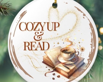 Puede incluir: Adorno navideño redondo y blanco con la frase "COZY UP & READ" en marrón. Presenta una ilustración de un libro abierto, una taza de café y un diseño en forma de corazón con detalles dorados. Colgando de un cordón dorado.