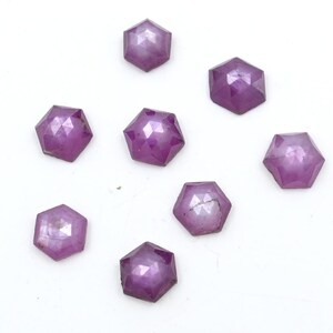 May include: Eight purple faceted gemstones, each with a hexagonal shape, are arranged on a white surface.