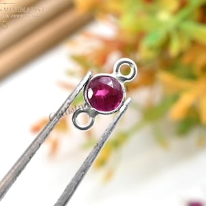 May include: A small, round, faceted deep pink gemstone connector set in a silver-toned metal frame with two circular loops on opposite sides. The piece is held by metal tweezers, showcasing the vibrant color and the reflective surface of the stone.