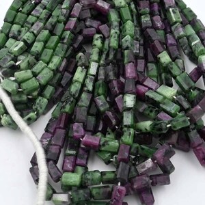 May include: Strands of rectangular beads in shades of green and purple. The beads are strung together on a white string, creating a textured and colorful display. Ideal for jewelry making.