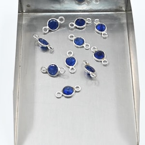 Puede incluir: Una colección de pequeñas piedras preciosas azules de forma ovalada engastadas en metal plateado, expuestas en una bandeja de acero inoxidable. Cada gema tiene un lazo plateado a cada lado. Las gemas son de un color azul intenso y vibrante.