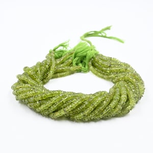 May include: A bundle of olive-green gemstone beads, strung together with a green tassel. The beads are small, round, and have a slightly translucent appearance. The beads are arranged in a circular pattern against a white background.