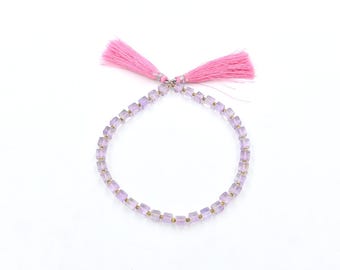 Può includere: Un delicato braccialetto con piccole perline quadrate viola chiaro, infilate con dettagli color oro. Il braccialetto è rifinito con un paio di nappe rosa acceso, che aggiungono un tocco di colore e un tocco di fantasia al design.