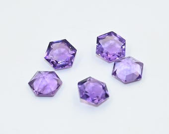 Könnte beinhalten: Fünf facettierte, sechseckige Amethyst-Edelsteine. Die Edelsteine haben eine tiefe lila Farbe und eine polierte, reflektierende Oberfläche. Die Edelsteine sind auf weißem Hintergrund angeordnet und zeigen ihre geometrische Form und Farbe.