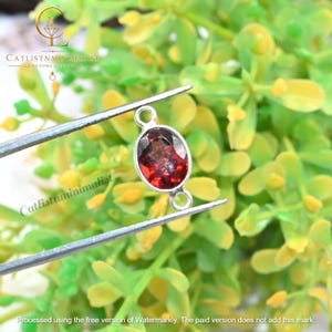 Pode incluir: Um conector de pedra preciosa granada vermelha de formato oval com superf&iacute;cie facetada, engastado em uma moldura de metal prateado com dois pequenos elos circulares nas extremidades, segurado por pin&ccedil;as sobre um fundo de folhagem verde.