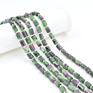 May include: A strand of green and pink gemstone beads, each bead is rectangular and faceted. The beads are strung on a white thread.