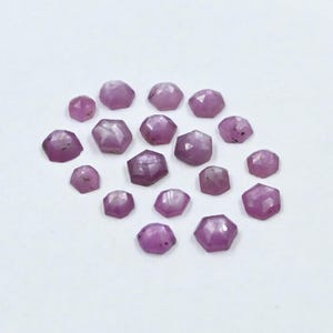 May include: A collection of 18 faceted pink gemstones, each with a unique shape and size. The stones are arranged on a white surface.