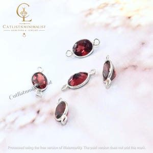 Pode incluir: Cinco pedras preciosas granada vermelho-escuras de formato oval, engastadas em arma&ccedil;&otilde;es de metal prateado. Cada pe&ccedil;a possui dois pequenos elos circulares nas extremidades opostas, projetados como conectores para joias.
