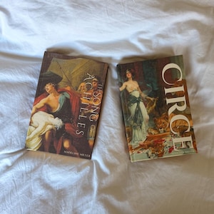 May include: Two hardback books with vibrant covers rest on a white surface. One book is titled "The Song of Achilles" and the other is titled "Circe". Both books showcase classical art illustrations.