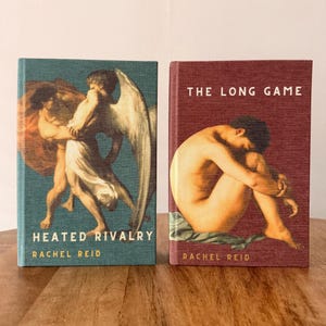 May include: Two hardcover books with artistic covers. The book on the left is teal with an image of two figures and the title "Heated Rivalry" by Rachel Reid. The book on the right is maroon with an image of a person and the title "The Long Game" by Rachel Reid.