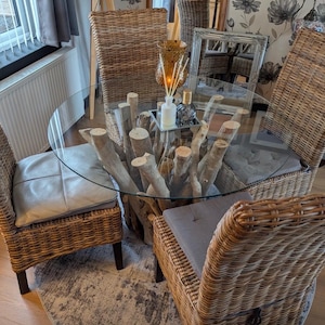 May include: A round glass-top dining table with a base constructed from multiple light-coloured driftwood branches. The table is surrounded by four high-backed woven wicker chairs, each featuring a square seat cushion in shades of grey.