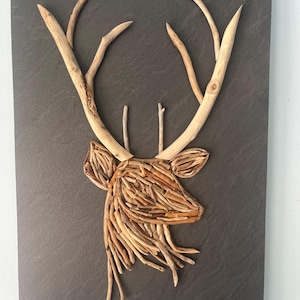 May include: A deer head made of driftwood, mounted on a dark gray slate background. The deer's antlers are made of long, thin pieces of driftwood, and its head is made of smaller, more intricate pieces of driftwood. The piece is signed "L. Martin" in the lower right corner.