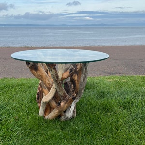 May include: A round glass-topped table with a base constructed from intertwined driftwood. The natural wood base supports the glass surface. The table is set on grass, with a beach and sea visible in the background.