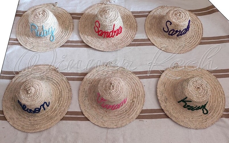 May include: Six straw hats with different names embroidered on them in various colors. The names are Ruby, Sandra, Sarah, Leeson, Brandon, and Krist.