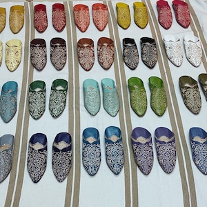 K&ouml;nnte beinhalten: Eine Auswahl an bunten Lederschuhen mit aufwendigen Stickereien. Die Schuhe sind in Reihen auf einer wei&szlig;en und braunen gestreiften Oberfl&auml;che angeordnet.