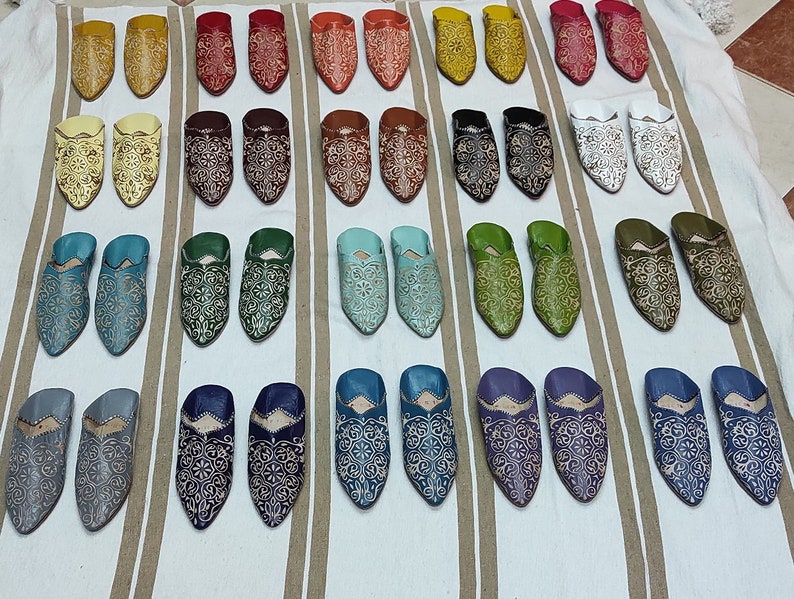 K&ouml;nnte beinhalten: Eine Auswahl an bunten Lederschuhen mit aufwendigen Stickereien. Die Schuhe sind in Reihen auf einer wei&szlig;en und braunen gestreiften Oberfl&auml;che angeordnet.