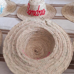 May include: A close-up of a straw hat with a red embroidered name "Sandra" on the front. The hat has a white pom-pom on top and is sitting on a striped fabric surface.