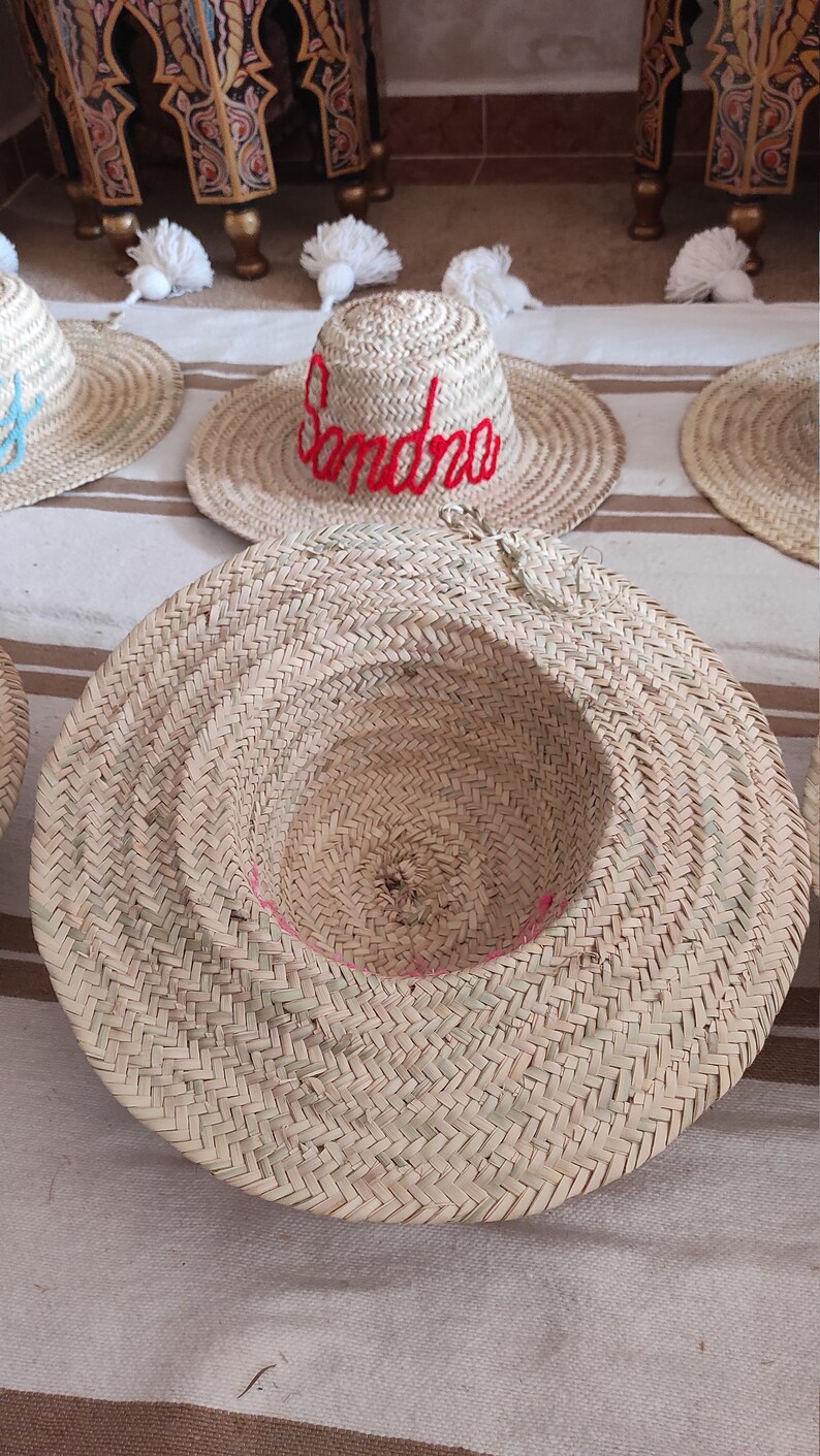 May include: A close-up of a straw hat with a red embroidered name "Sandra" on the front. The hat has a white pom-pom on top and is sitting on a striped fabric surface.