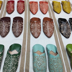 K&ouml;nnte beinhalten: Eine Auswahl an bunten Lederschuhen mit aufwendigen Stickereien. Die Schuhe sind in Reihen auf einem gestreiften Stoffhintergrund angeordnet.