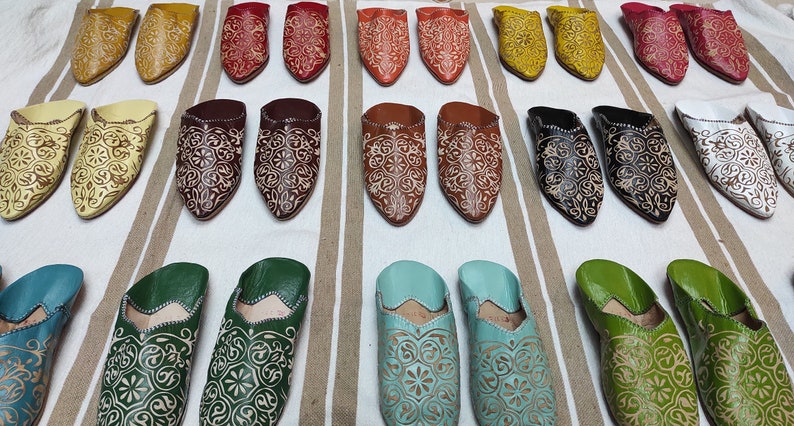 K&ouml;nnte beinhalten: Eine Auswahl an bunten Lederschuhen mit aufwendigen Stickereien. Die Schuhe sind in Reihen auf einem gestreiften Stoffhintergrund angeordnet.
