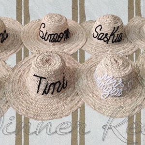 May include: Nine straw hats with different names embroidered on them in black thread. The hats are arranged in a semi-circle on a striped background.