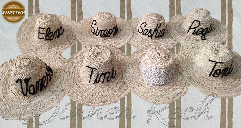 May include: Nine straw hats with different names embroidered on them in black thread. The hats are arranged in a semi-circle on a striped background.