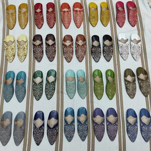 K&ouml;nnte beinhalten: Eine Sammlung von 24 Paar Lederschlappen in verschiedenen Farben, darunter Gelb, Rot, Braun, Blau, Gr&uuml;n, Wei&szlig; und Grau. Jeder Schlappen ist mit aufwendigen Blumenstickereien verziert.