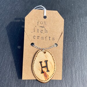 May include: A wooden pendant necklace with the letter H burned into it. The pendant is oval shaped and has a silver chain. The necklace is on a brown paper tag with the text "fox itch crafts" printed on it.