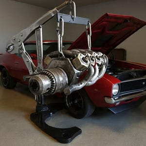 May include: A sculpture crafted from repurposed metal tools and automotive parts, featuring a large silver locking wrench and a drill chuck suspended by chains from a metal frame, all mounted on a solid black base, positioned before a red car.