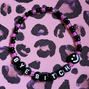 May include: A black beaded bracelet with pink beads and black letter beads spelling "BYE BITCH" with a black smiley face bead. The bracelet is on a pink and black leopard print background.