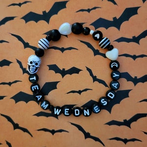 May include: A black and white beaded bracelet with the words "Team Wednesday" and "Day" spelled out. The bracelet also features a skull charm, hearts, and black and white striped beads.