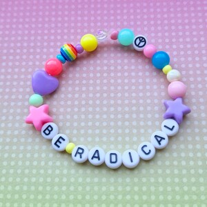 Pode incluir: Um bracelete de contas coloridas com as palavras "BE RADICAL" escritas em contas brancas com letras pretas. O bracelete também inclui um amuleto de sinal de paz, estrelas e corações.