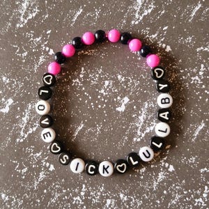 May include: A bracelet made of black, white, and pink beads. The bracelet spells out "LOVE SICK LULLABY" using white letter beads and heart-shaped beads. The bracelet is displayed on a grey speckled surface.