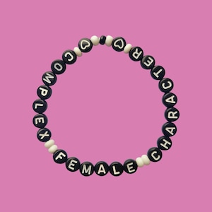 Puede incluir: Una pulsera de cuentas negras y blancas que deletrean "COMPLEX FEMALE CHARACTER". La pulsera presenta cuentas en forma de corazón y está sobre un fondo rosa. Un accesorio único.