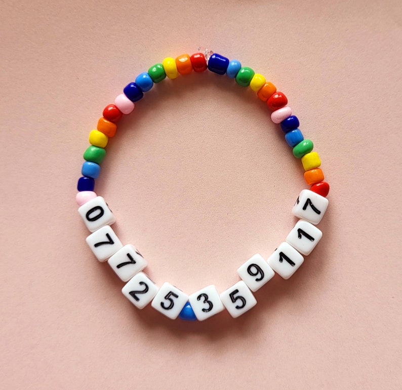 May include: A rainbow beaded bracelet with white beads that have numbers on them. The numbers on the beads are 07725359117.