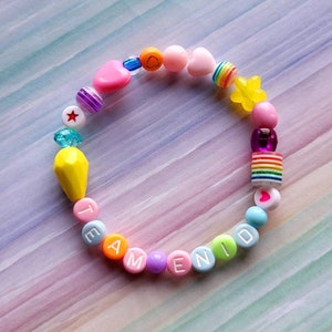 May include: A colourful beaded bracelet with the word "TEAM" and "ENID" spelled out in white letters. The bracelet also features a yellow star, a rainbow bead, a pink heart, and other colourful beads.