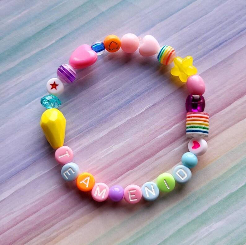 May include: A colourful beaded bracelet with the word "TEAM" and "ENID" spelled out in white letters. The bracelet also features a yellow star, a rainbow bead, a pink heart, and other colourful beads.