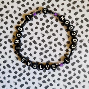 May include: A black beaded bracelet with white letter beads spelling "I DON'T NO COCO I EVOLVE". The bracelet includes purple accent beads and a crescent moon bead. The background is a grey and white dotted pattern.