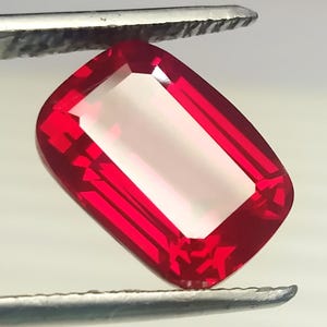 Puede incluir: Una gema facetada de forma rectangular con un vibrante color rojo. La gema está sujeta por una herramienta plateada, mostrando sus facetas pulidas y su claridad interna. Las esquinas son redondeadas y el centro es un tono más claro de rojo.