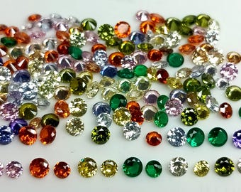 May include: Assortment of colorful round gemstones in various shades of green, blue, purple, pink, orange, and yellow. The gemstones are arranged in a loose pile on a white surface.